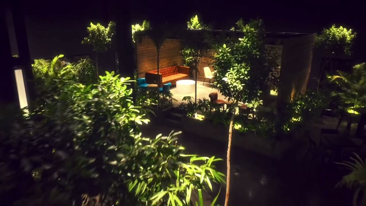 A tufted sofa, teal armchairs and a round white table under a pergola, lit among the trees at night